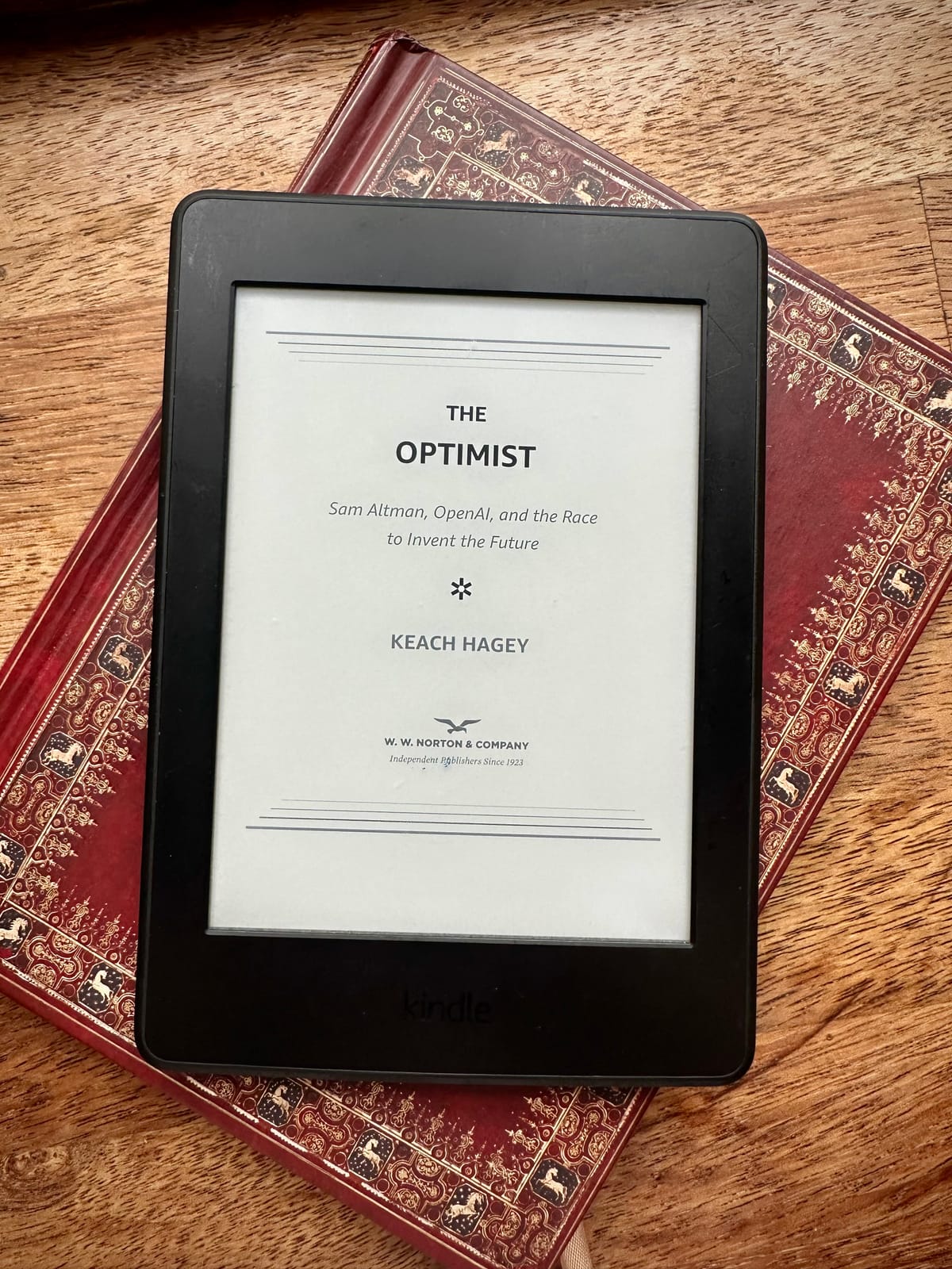 A kindle on top of a notebook. The Kindle shows the cover of The Optimist.